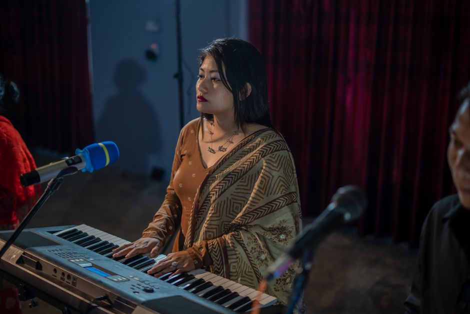 A woman playing a keyboard on stage with a microphone, wearing a saree.